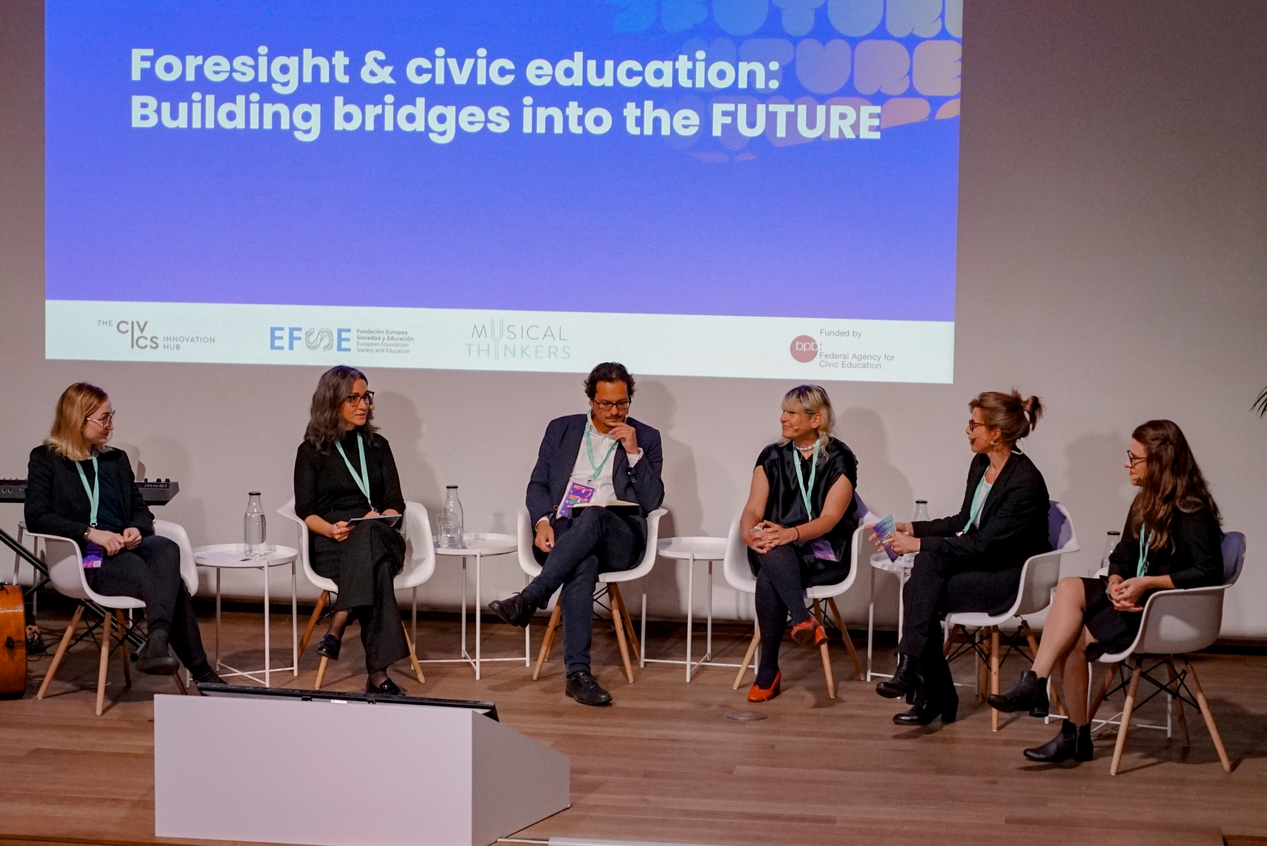 A panel of six people sits on stage discussing "Foresight & civic education: Building bridges into the FUTURE" with a screen backdrop.